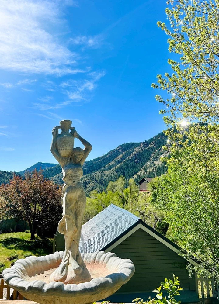 A stone statue of a woman holding a large urn above her head in a garden  at Joytown, a transformational retreat & tantra sanctuary in Colorado.