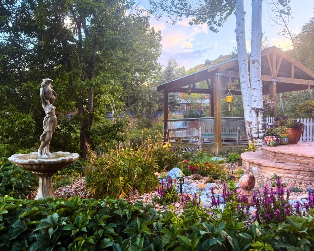 A lush garden with a decorative statue of a woman holding a vessel on a fountain, surrounded by colorful flowers, trees, and a wooden gazebo in the background at Joytown, a transformational retreat & tantra sanctuary in Colorado.