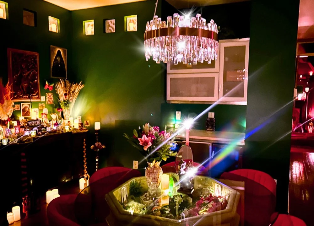 Interior of a dimly lit room decorated with candles, flowers, and artwork on dark green walls, featuring a chandelier hanging above a table with a floral centerpiece at Joytown, a transformational retreat & tantra sanctuary in Colorado.