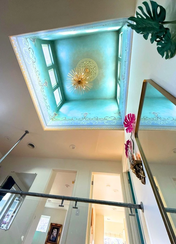 View of an ornate ceiling with a chandelier, four windows, and decorative borders, taken from the ground looking upward at Joytown, a transformational retreat & tantra sanctuary in Colorado.