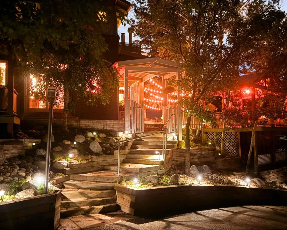 Nighttime view of a backyard with illuminated wooden stairs leading to a deck. The area is decorated with string lights and small ground lights, surrounded by trees and rocks at Joytown, a transformational retreat & tantra sanctuary in Colorado.