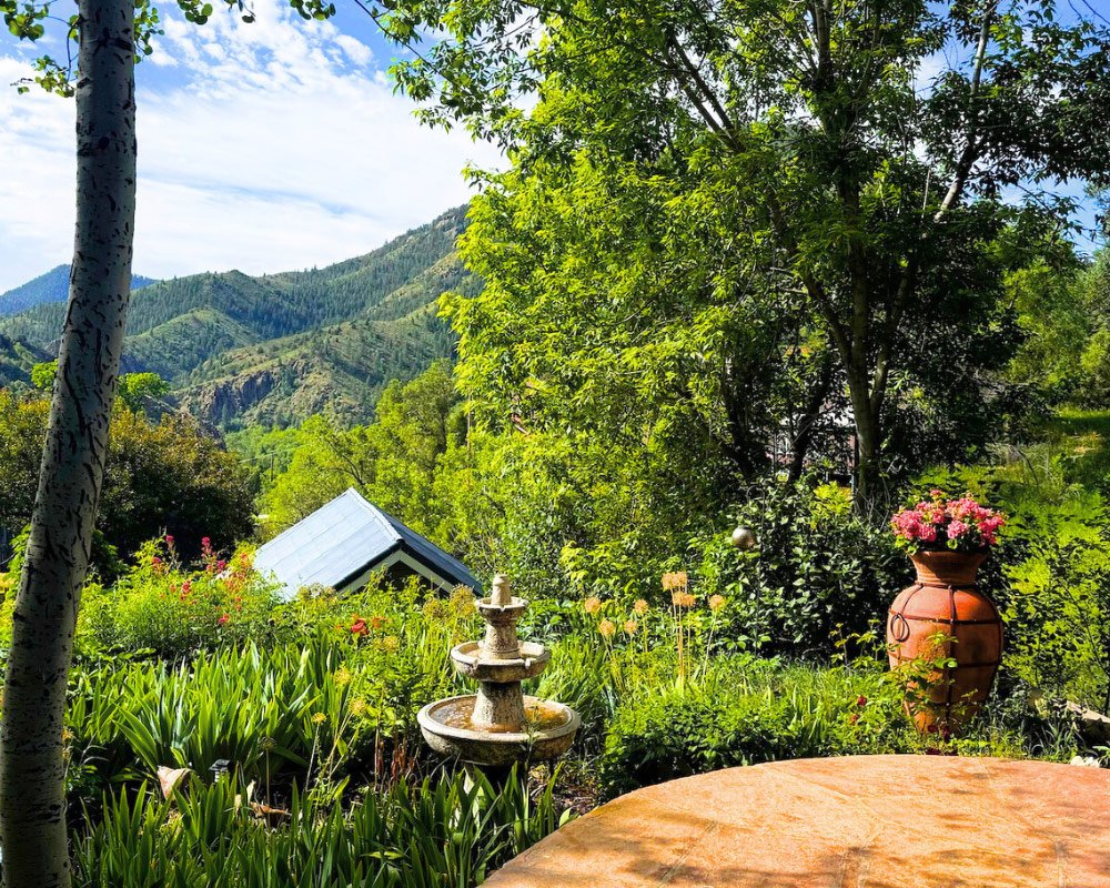 A lush garden scene with green plants, trees, and a background of mountain hills. There is a small fountain, a clay urn with pink flowers, and a table in the foreground at Joytown, a transformational retreat & tantra sanctuary in Colorado.