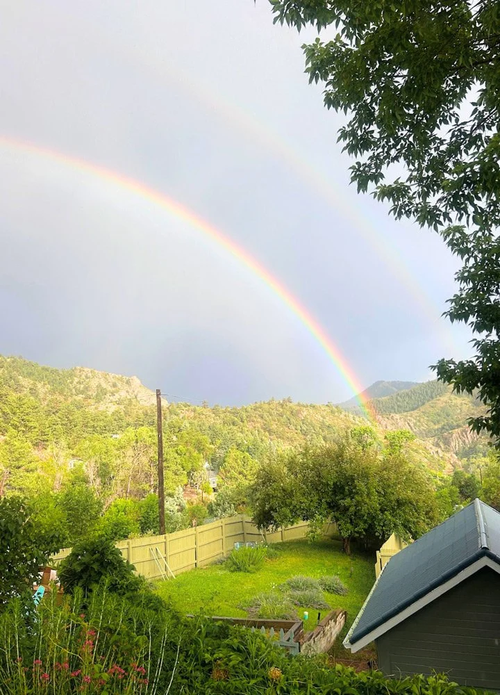 A backyard scene with a rainbow and a faint second rainbow in the sky at Joytown, a transformational retreat & tantra sanctuary in Colorado.