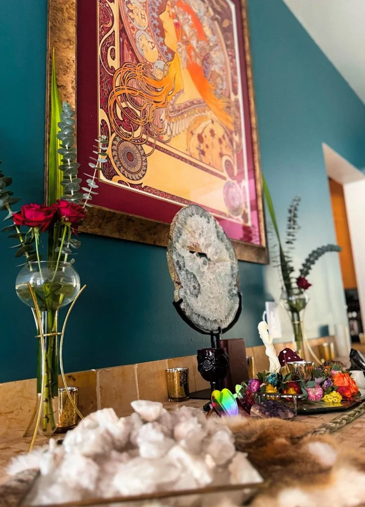 Decorative display with vases of flowers, a geode on a stand, and a tray of cotton and other items on a countertop against a colorful framed artwork and teal wall at Joytown, a transformational retreat & tantra sanctuary in Colorado.