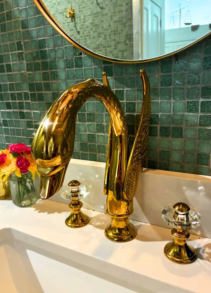 Close-up of a gold swan-shaped faucet on a white bathroom sink, with a bouquet of pink and yellow flowers in a glass jar and a mirror  at Joytown, a transformational retreat & tantra sanctuary in Colorado.