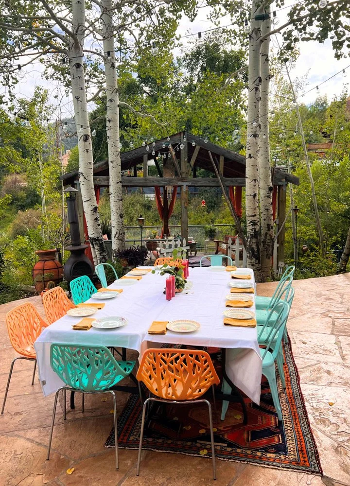 Outdoor dining setup on a stone patio, with a long rectangular table covered with a white tablecloth, colorful chairs around it, and a gazebo  at Joytown, a transformational retreat & tantra sanctuary in Colorado.