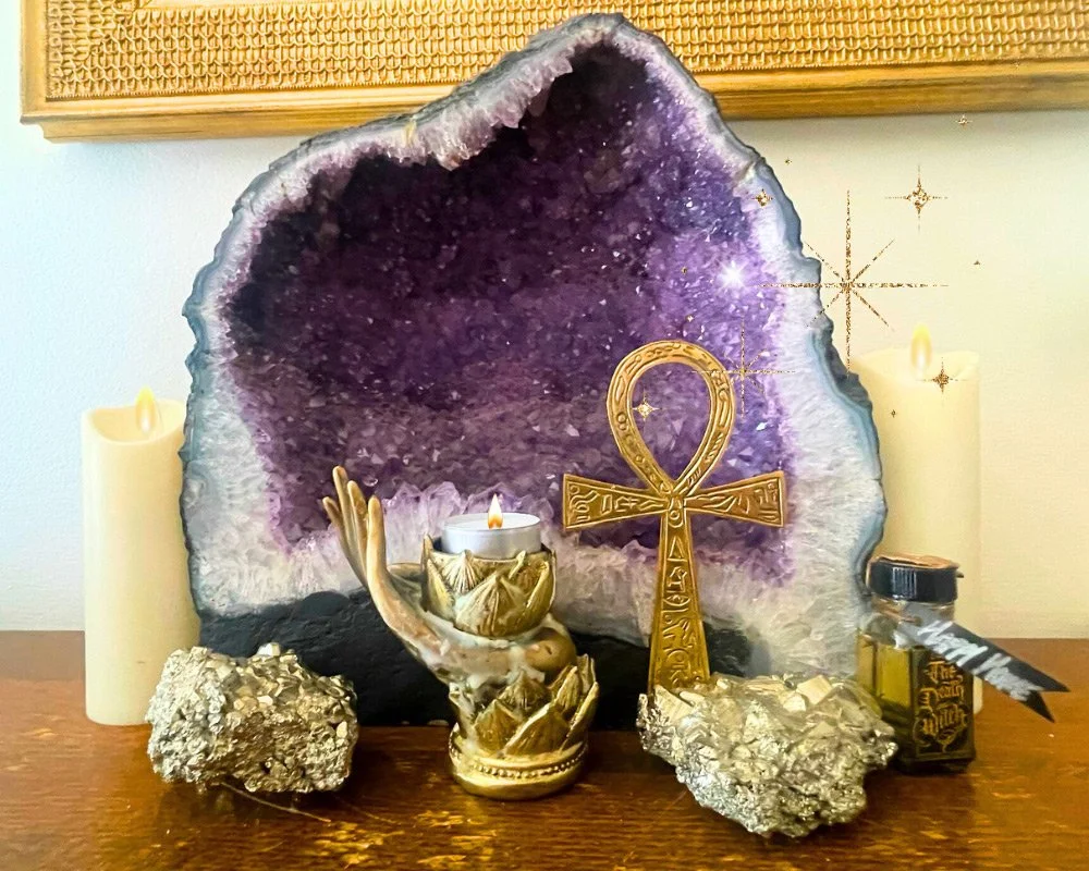 An altar with a large amethyst geode, a gold ankh symbol, a gold hand holding a candle, other smaller rocks, candles, and bottles at Joytown, a transformational retreat & tantra sanctuary in Colorado.
