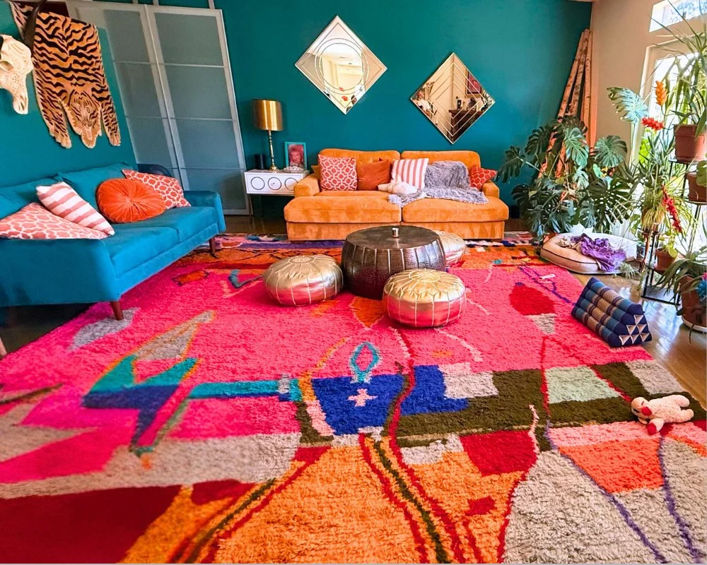 Colorful living room with teal and orange couches, patterned pillows, an abstract vibrant area rug, large plants near windows, and decorative wall mirrors at Joytown, a transformational retreat & tantra sanctuary in Colorado.