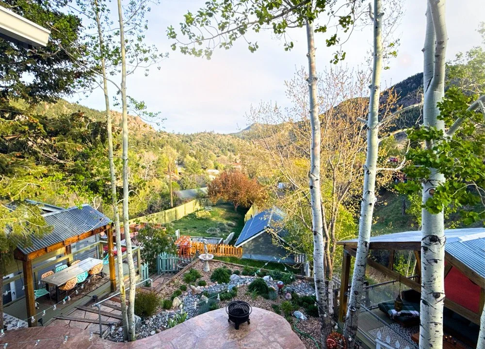 A backyard with a patio, trees, and a view of mountains in the distance, featuring a garden, outdoor seating, and a shed at Joytown, a transformational retreat & tantra sanctuary in Colorado.