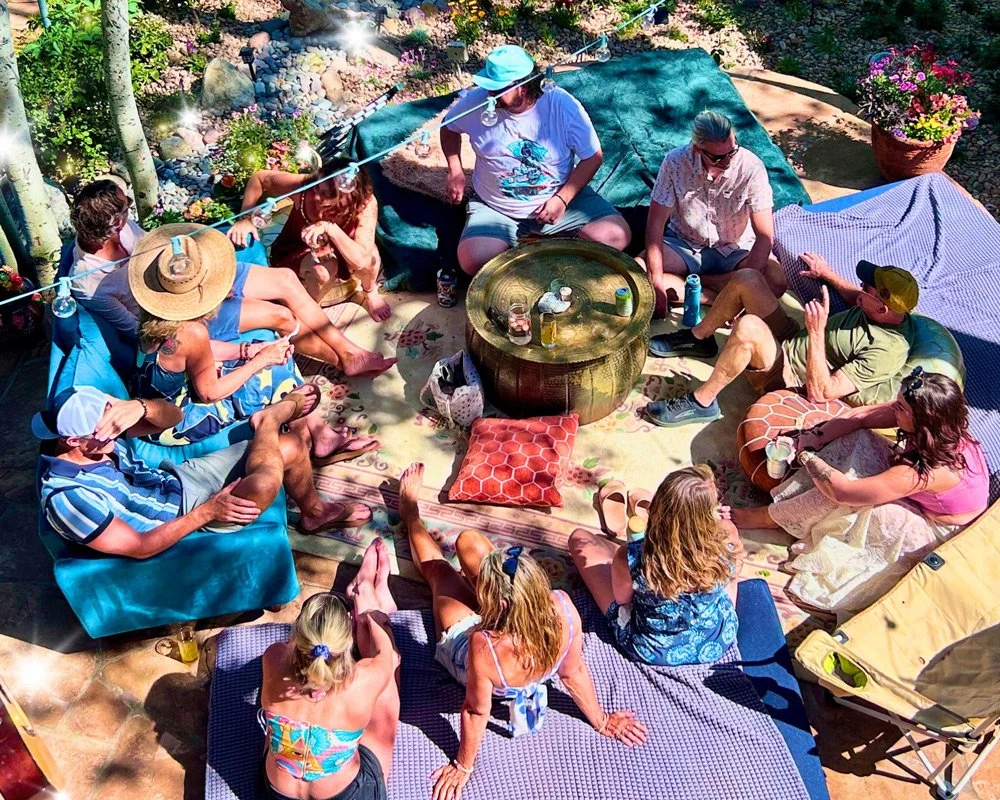 A group of people sitting in a circle outdoors on a sunny day, enjoying drinks and conversation, with colorful cushions and blankets on the ground at Joytown, a transformational retreat & tantra sanctuary in Colorado.