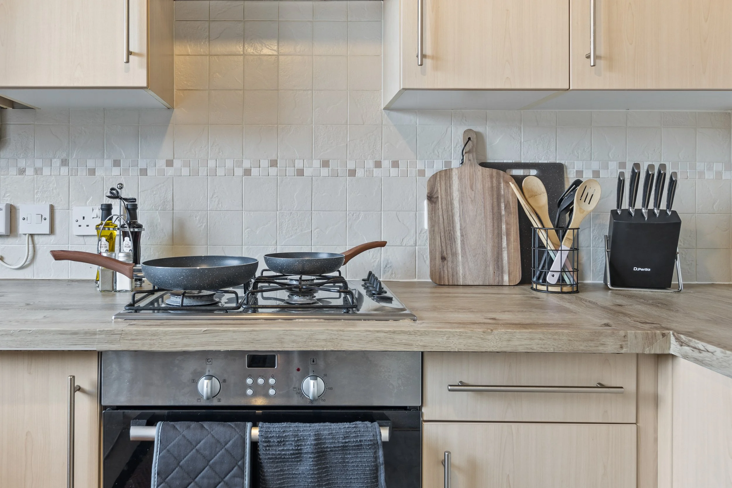 A modern kitchen with a gas stove, wooden cutting boards, and various utensils like spatulas and knives organized in containers. Light-colored cabinetry and tiled backsplash are visible.