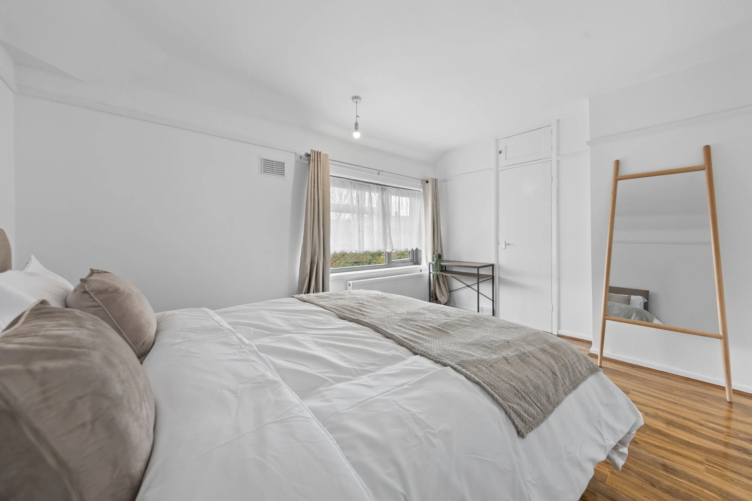 A minimalist bedroom with a large bed covered in white bedding and beige pillows. There are beige curtains on the window, a small black metal shelf with a plant, and a full-length wooden mirror. The room has white walls, a wooden floor, and a simple ceiling with a single exposed light bulb.