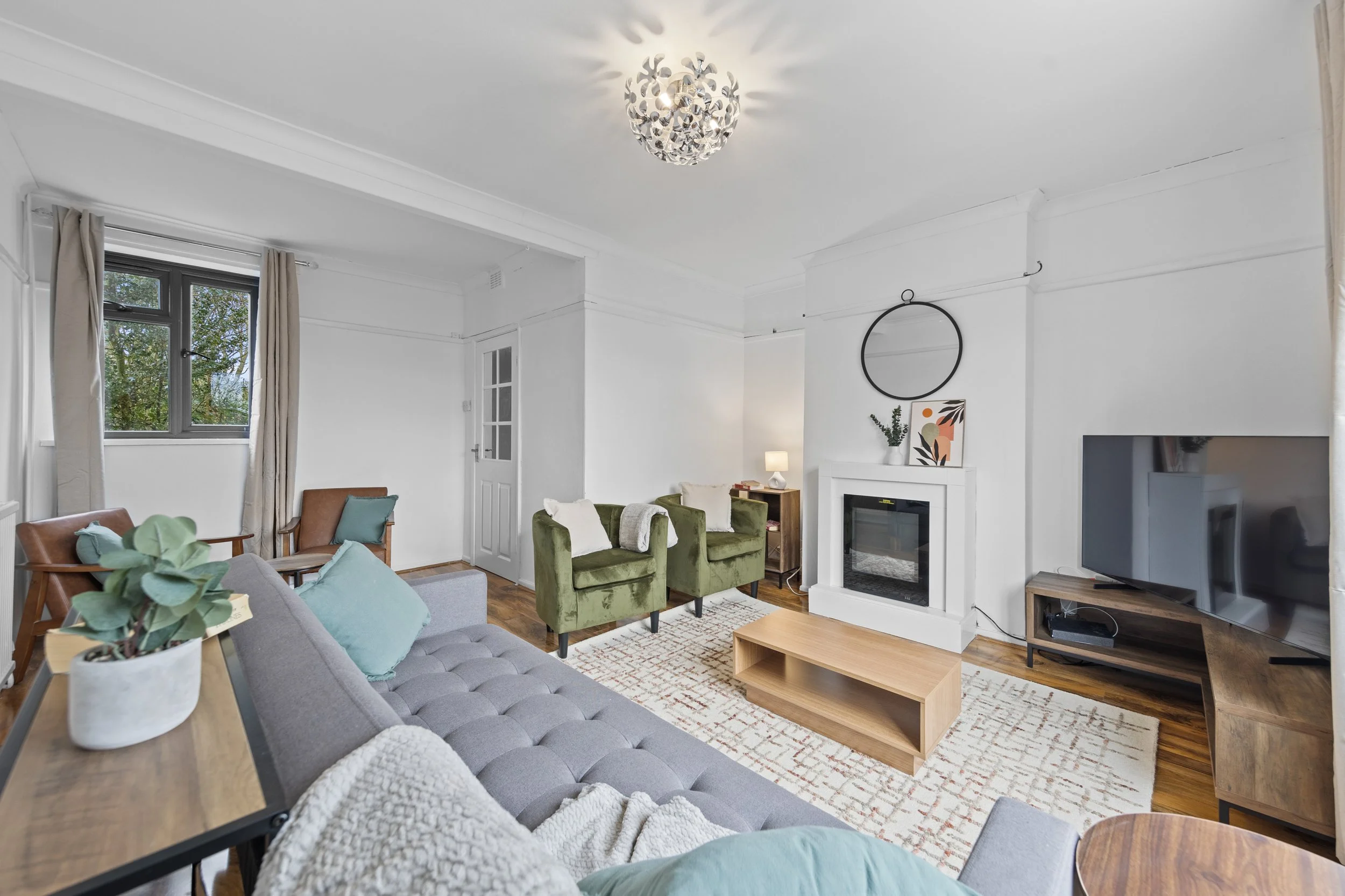 A living room with a gray tufted sofa, green armchairs, a wooden coffee table, a TV, a fireplace, windows with beige curtains, and modern decor including a round mirror and artwork.