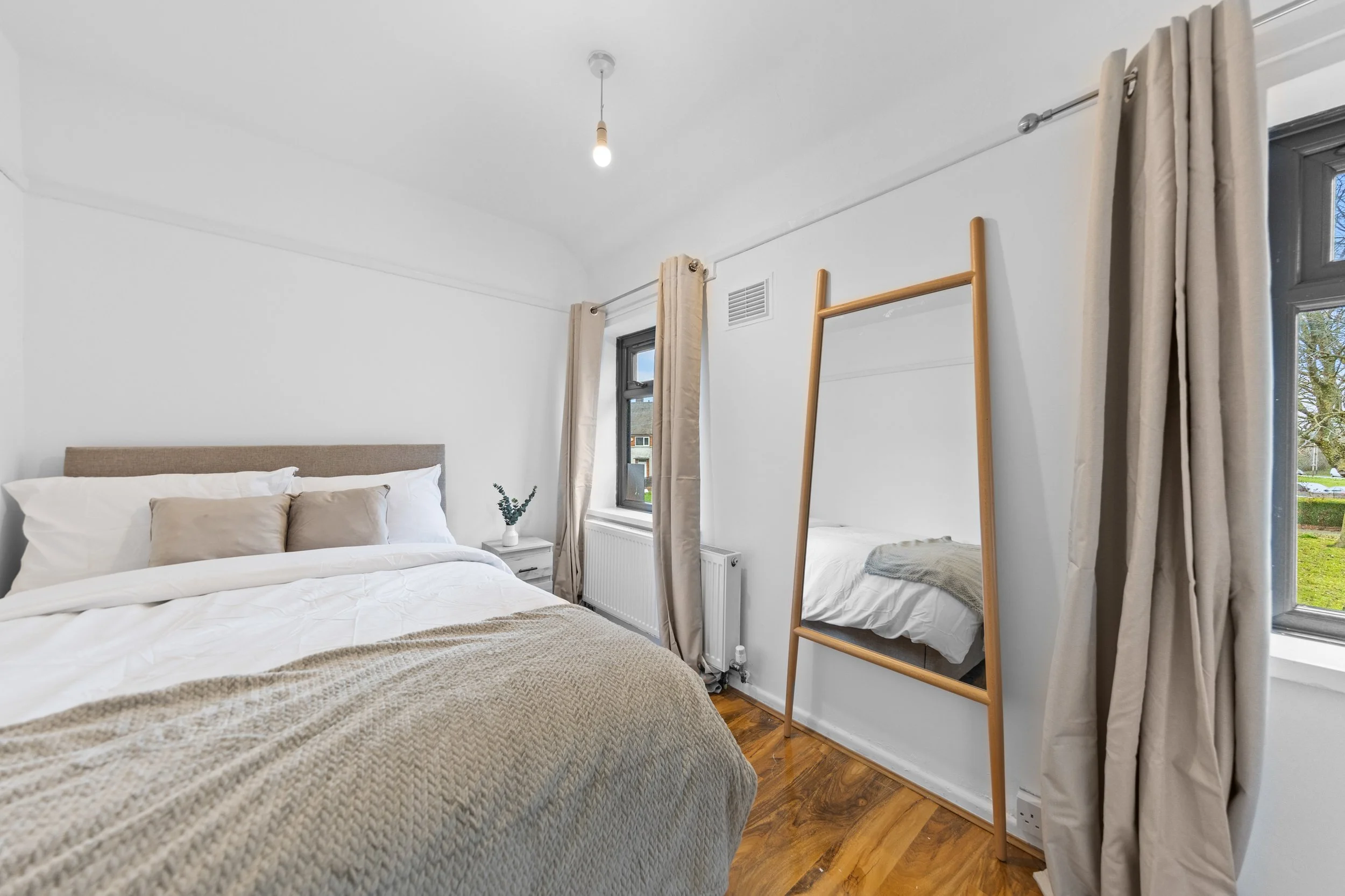 A bedroom with two beds, beige and white bedding, a window with beige curtains, a wooden ladder against the wall, and a small white bedside table with a plant. Natural light from outside illuminates the room.