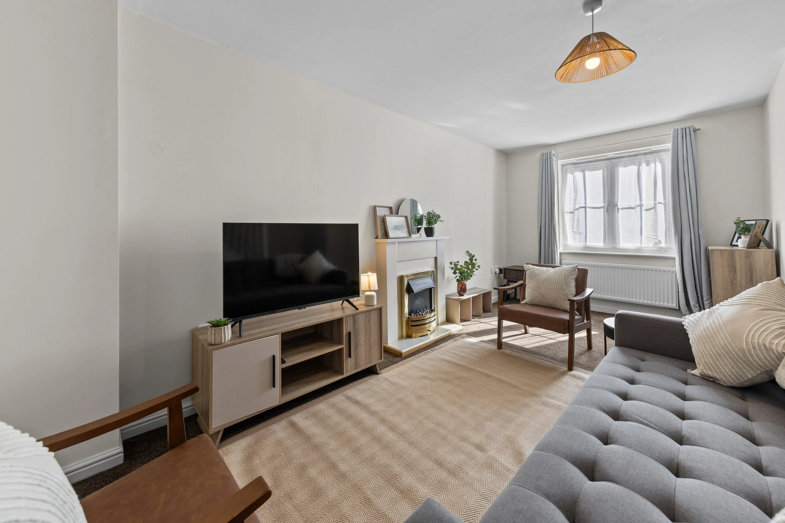 Living room with a large window, white walls, a gray tufted sofa, a brown armchair, a wooden media console with a TV, plants, artwork, and a fireplace with a mirror above.