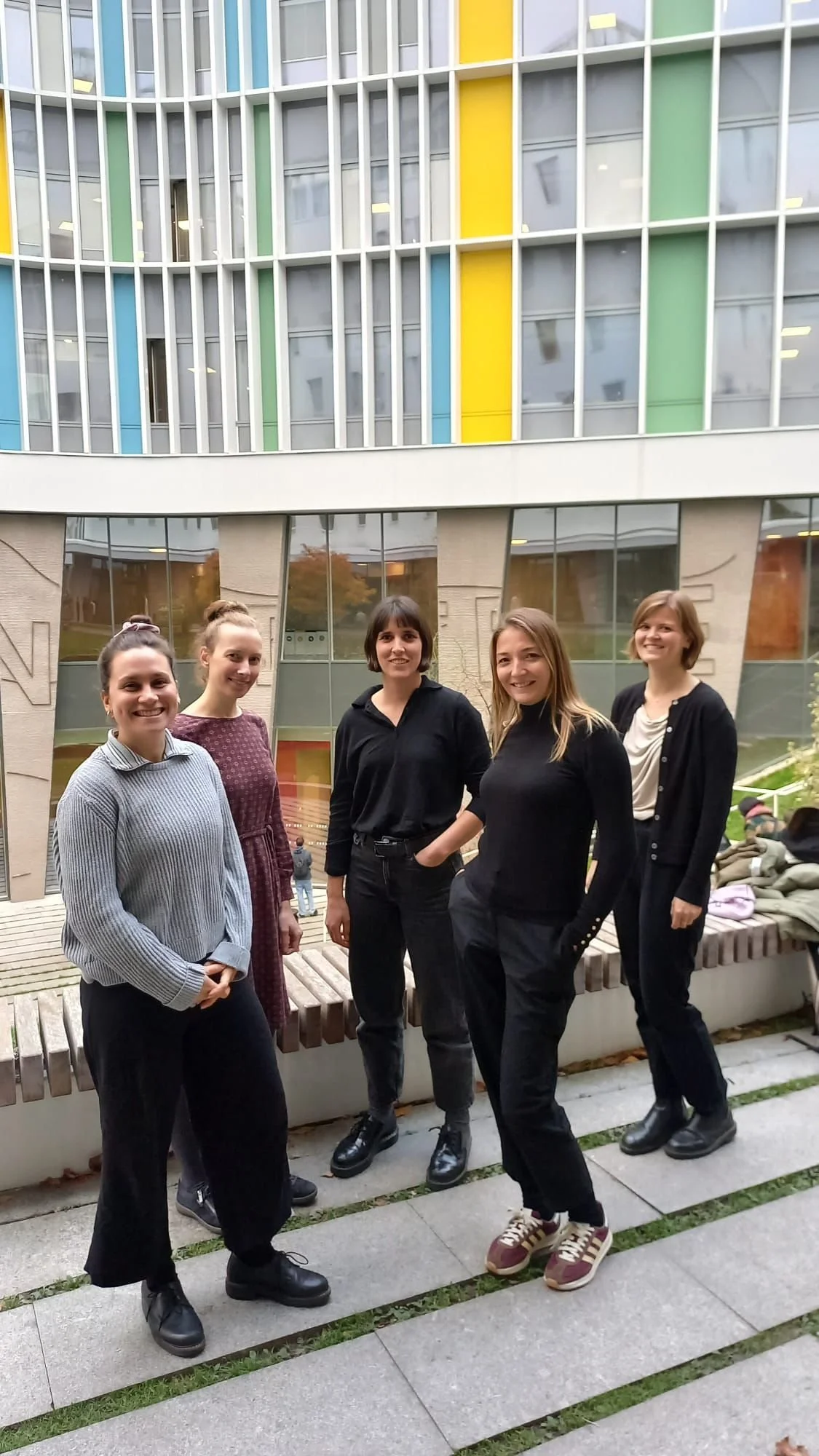 Fünf Personen stehen in einer Reihe im Freien vor einem Gebäudekomplex mit mehreren Etagen und Balkonen, bei bewölktem Himmel.