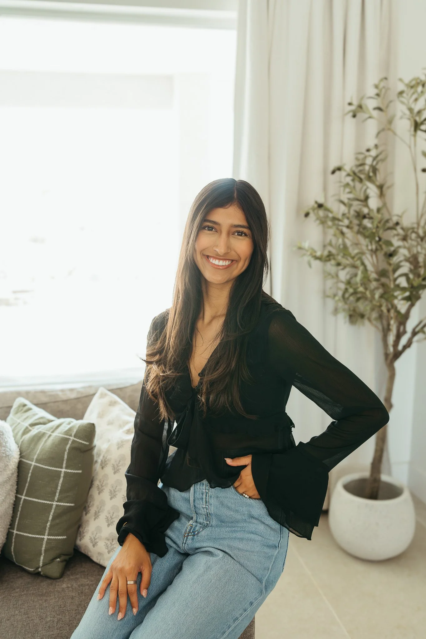 A woman with long dark hair smiling while sitting on a couch in a well-lit room, with decorative pillows and a potted plant nearby.