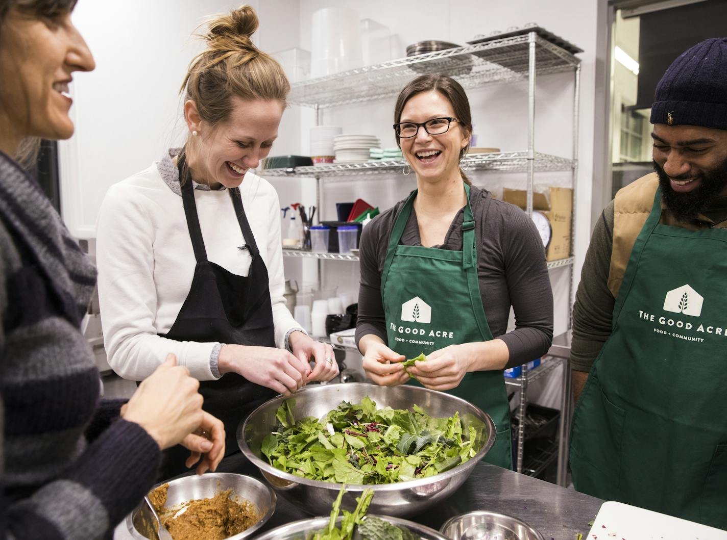 Three women and a man laughing while preparing a large salad in a professional kitchen coop.