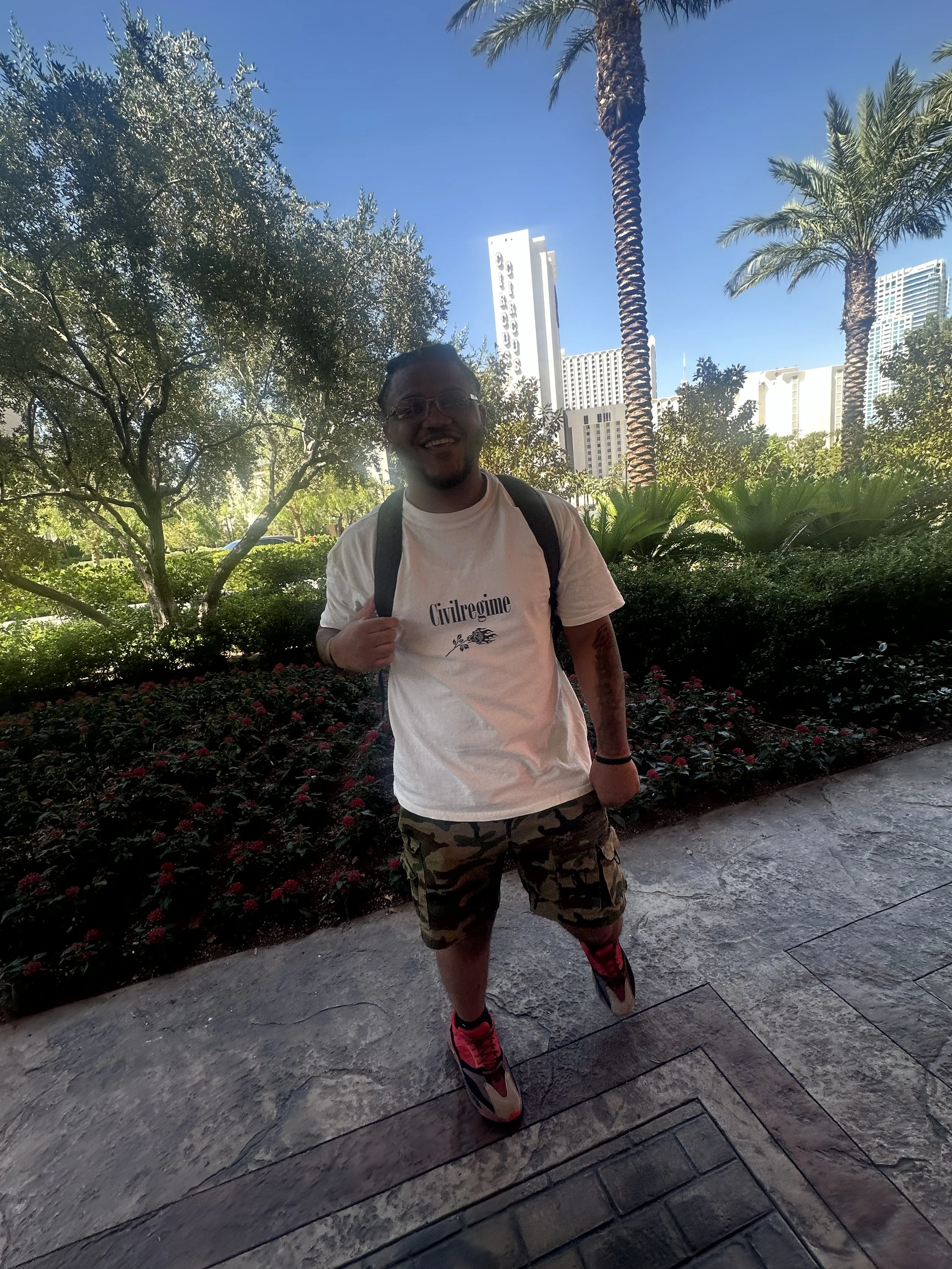 Smiling man standing outdoors in a garden with palm trees and tall buildings in the background, wearing a white t-shirt with 'Civil Regime' written on it, camouflage shorts, and colorful sneakers.