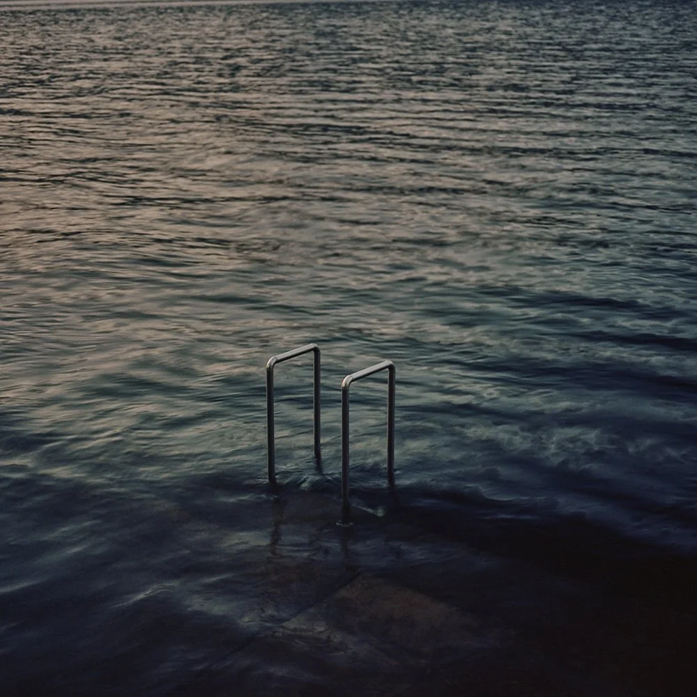 Una scaletta di metallo che si immerge in un lago o nel mare durante il tramonto, con acqua calma e superficie leggermente ondulata.