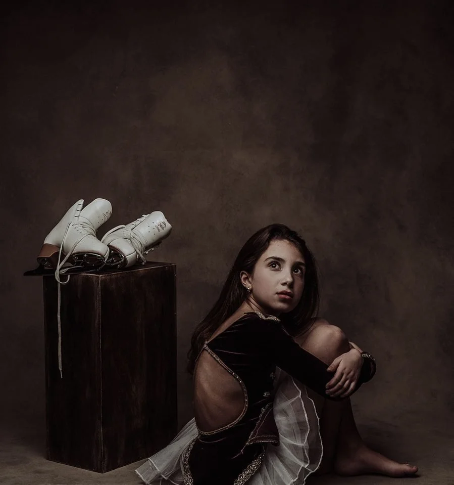Una ragazza seduta sul pavimento con le ginocchia piegate, vestita con un abito nero con schiena scoperta e un tutù bianco, in un ambiente scuro e neutro. Nella parte sinistra dell'immagine, su un supporto di legno scuro, ci sono un paio di pattini d