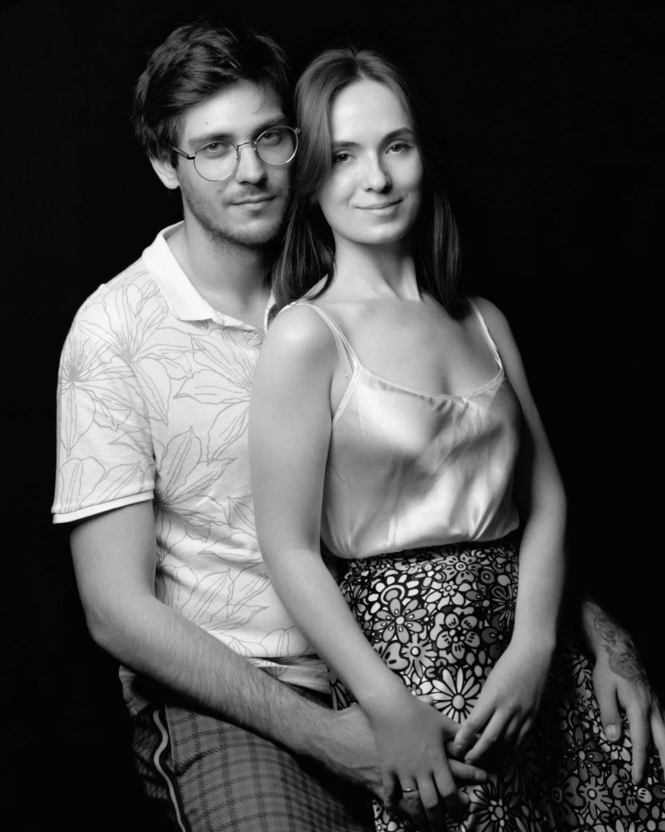 Ritratto di Una coppia di giovani adulti seduti uno accanto all'altro, lei con una gonna con un motivo floreale e lui con un maglione con disegni botanici. Entrambi sorridono leggermente, sfondo nero, in bianco e nero.