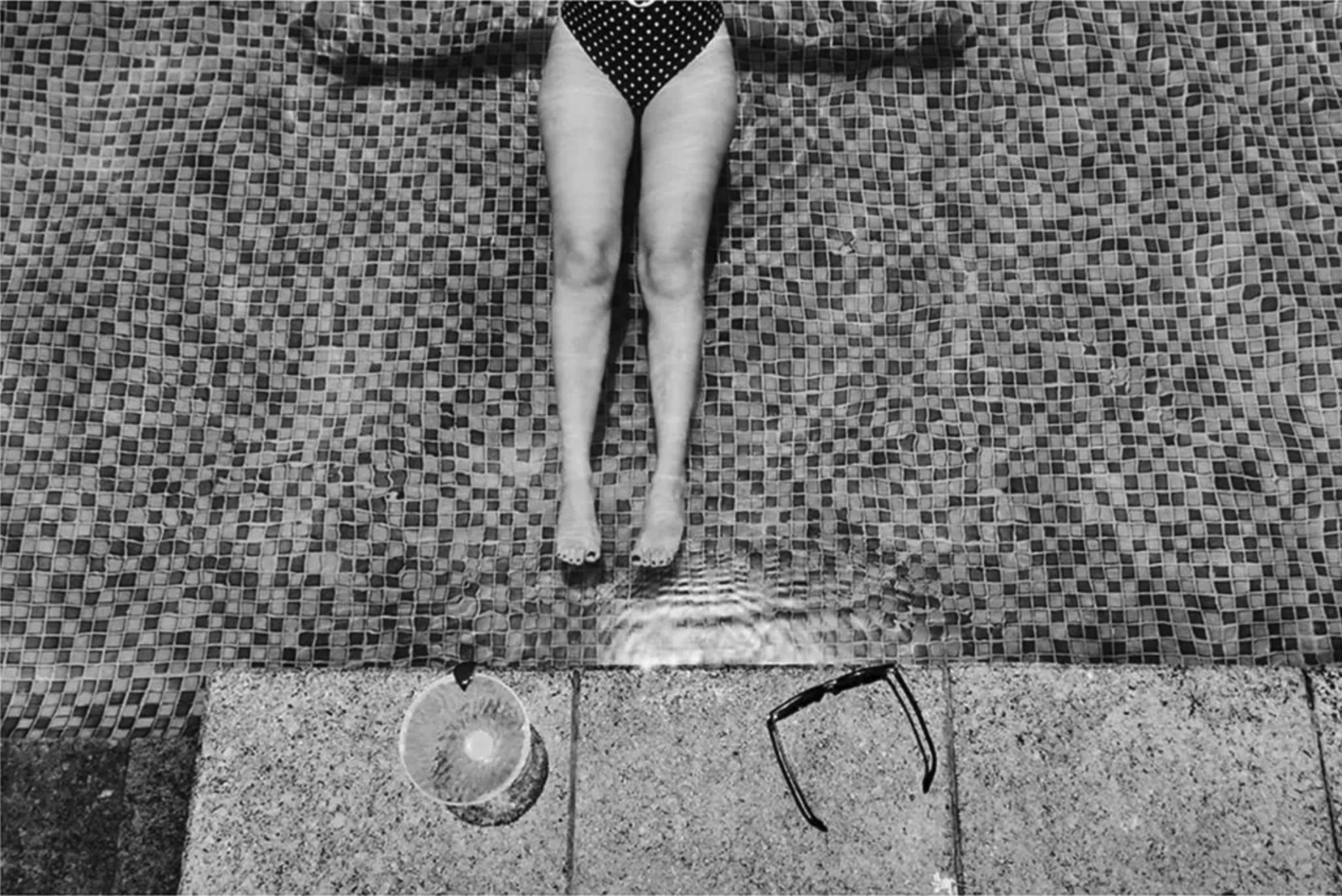 Una persona in bikini che sta sdraiata in una piscina con mosaici, con occhiali da sole sul pavimento e una bottiglia d'acqua vicino.