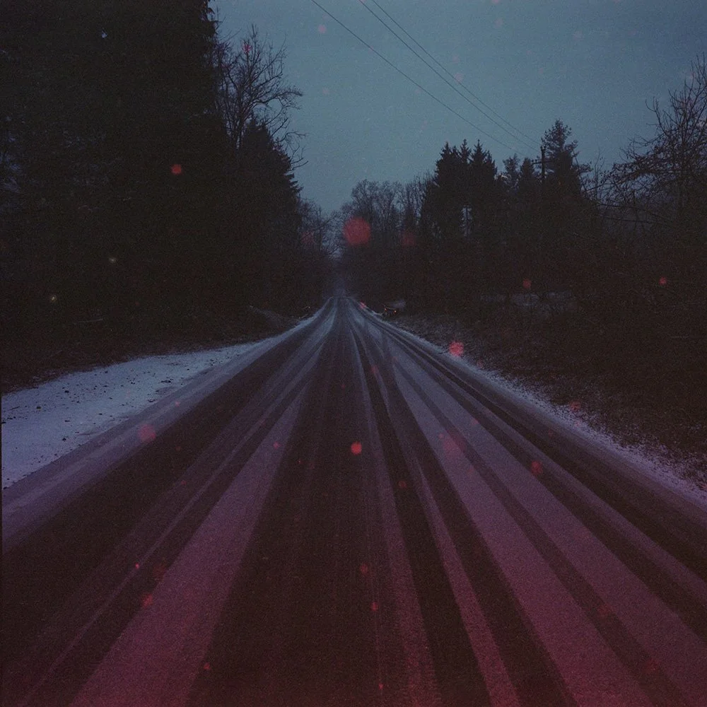 Strada di campagna bagnata, con neve e alberi ai lati, al tramonto o all'alba, con alcune luci rosse di pioggia o neve sul lente