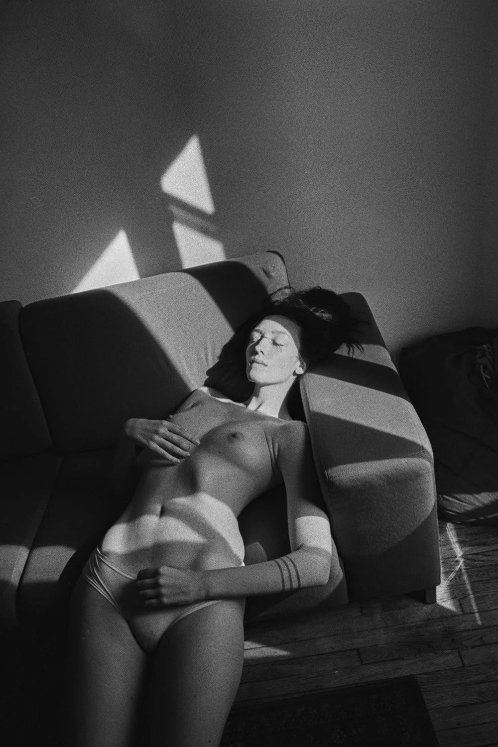 Donna sdraiata nuda su un divano in una stanza, con luci e ombre sulla parete.