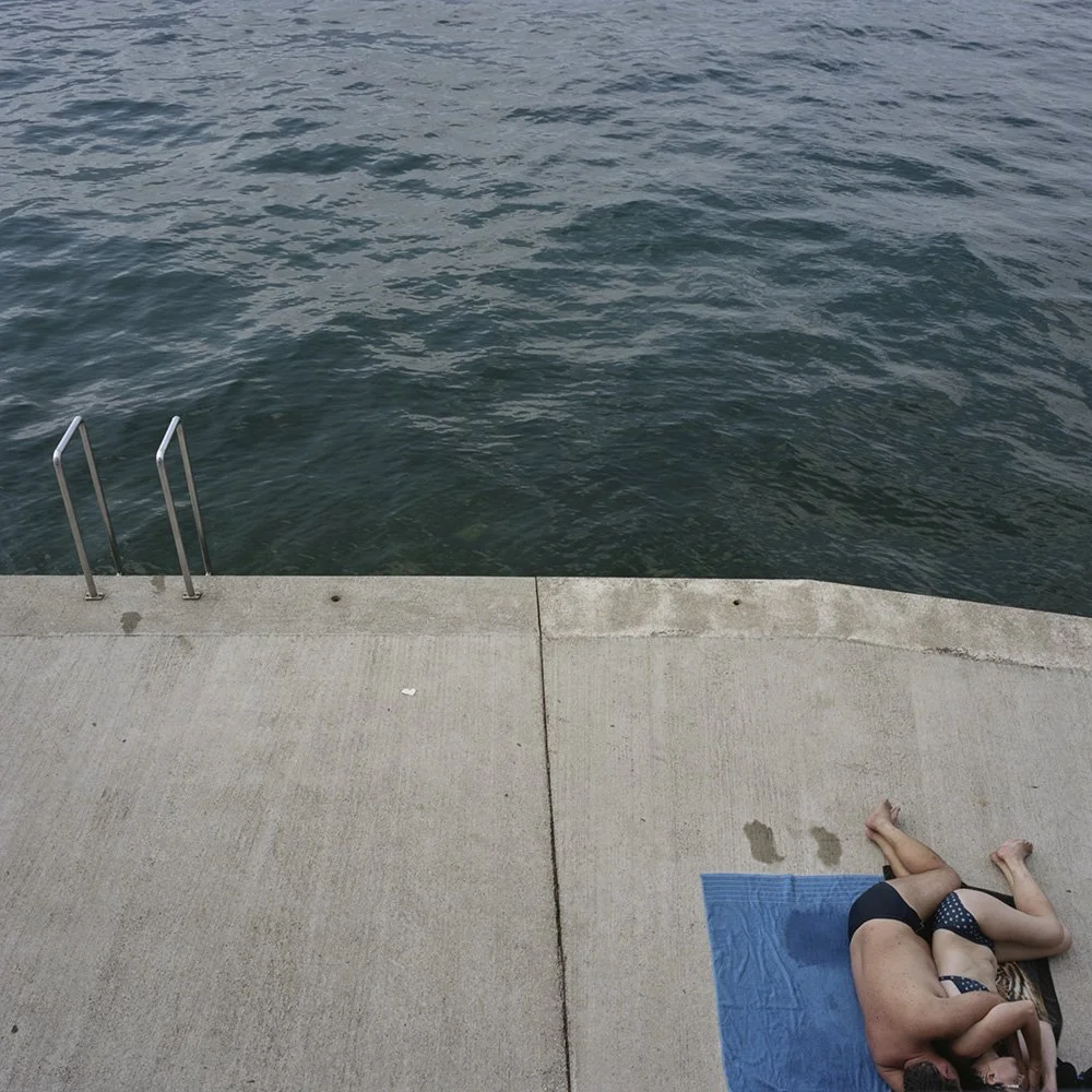 Donna sdraiata su un tappetino sulla banchina di un porto, in posizione di yoga o meditazione, con la parte superiore del corpo piegata in avanti.