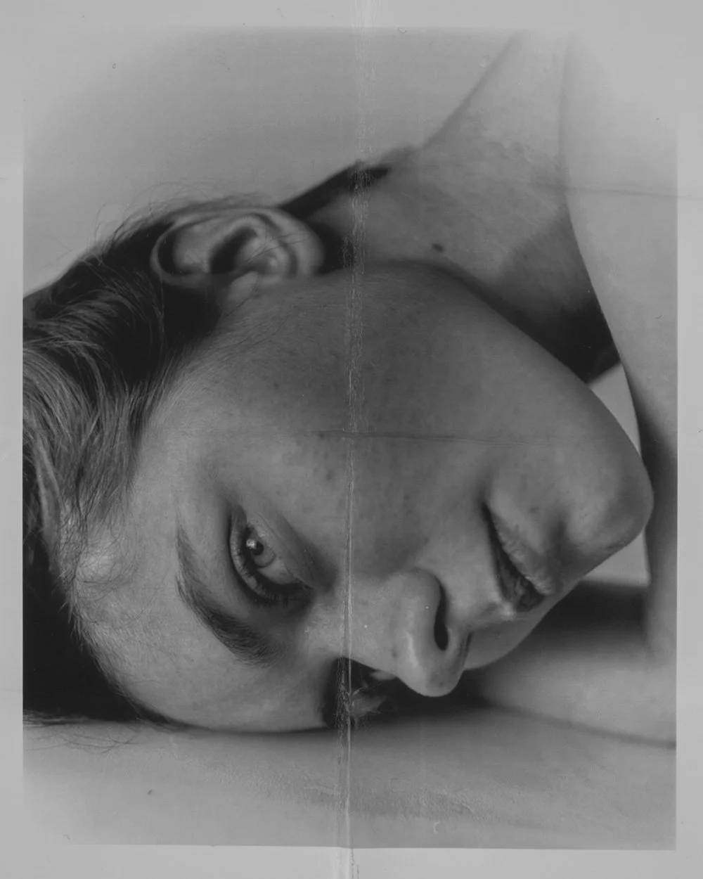 Foto in bianco e nero di una donna sdraiata, con il volto appoggiato su una superficie, occhi aperti e sguardo intenso.