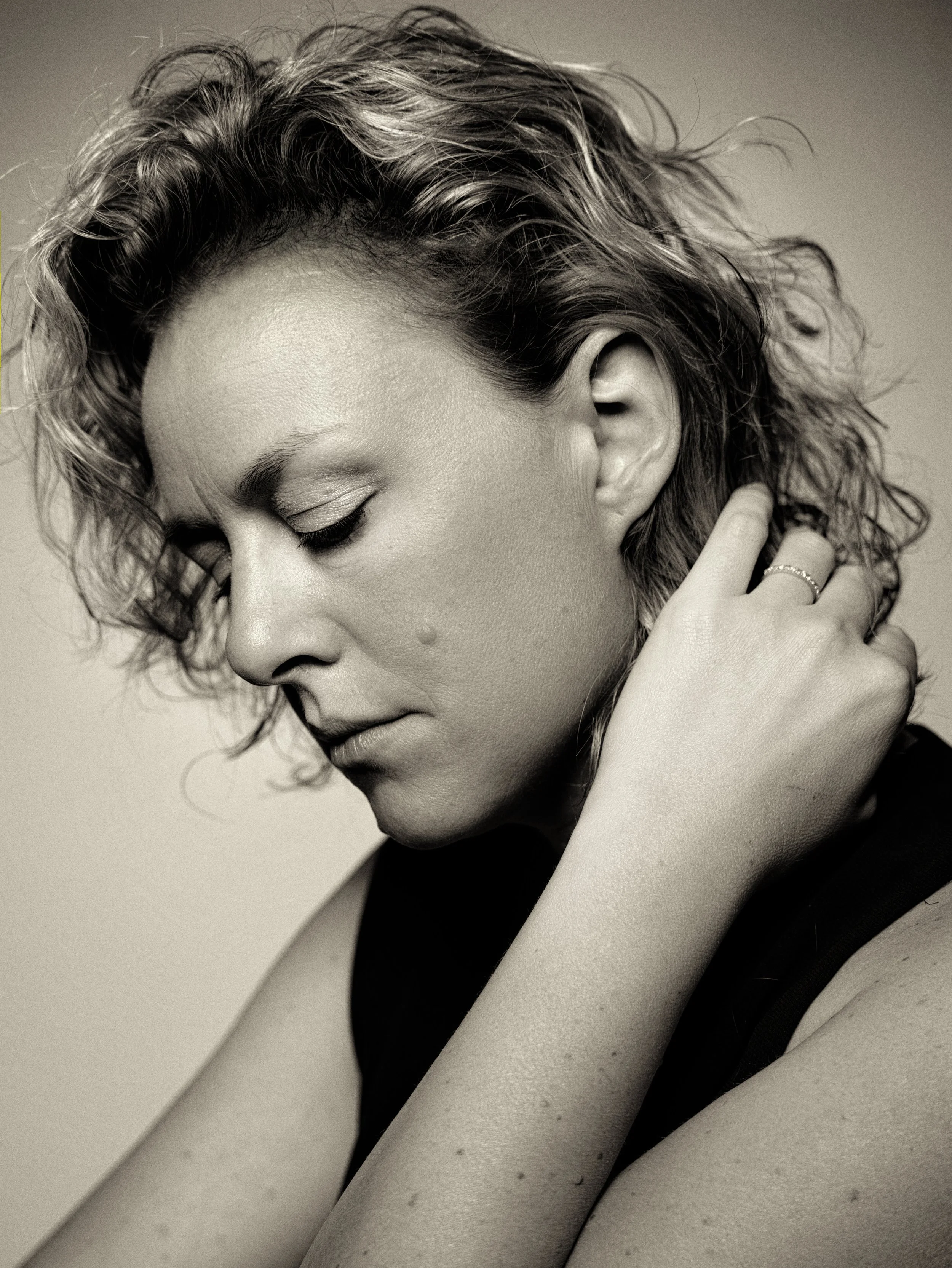 Ritratto in bianco e nero di una donna con i capelli ricci, occhi chiusi, che si sfiora la nuca con una mano, con alcuni nei visibili sulla pelle.