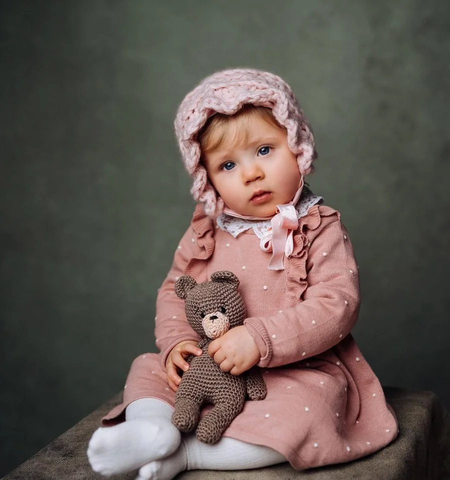 Una bambina con cappello di lana rosa, vestito rosa con dettagli in pizzo, che tiene un orsacchiotto marrone fatta a maglia, seduta su una superficie contro uno sfondo sfumato grigio-verde.