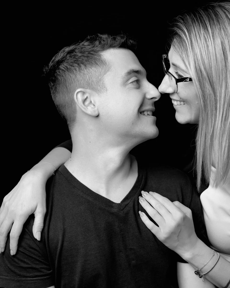 Una coppia di giovani in un abbraccio sorridenti, con il viso molto vicino, in una foto in bianco e nero su sfondo nero.