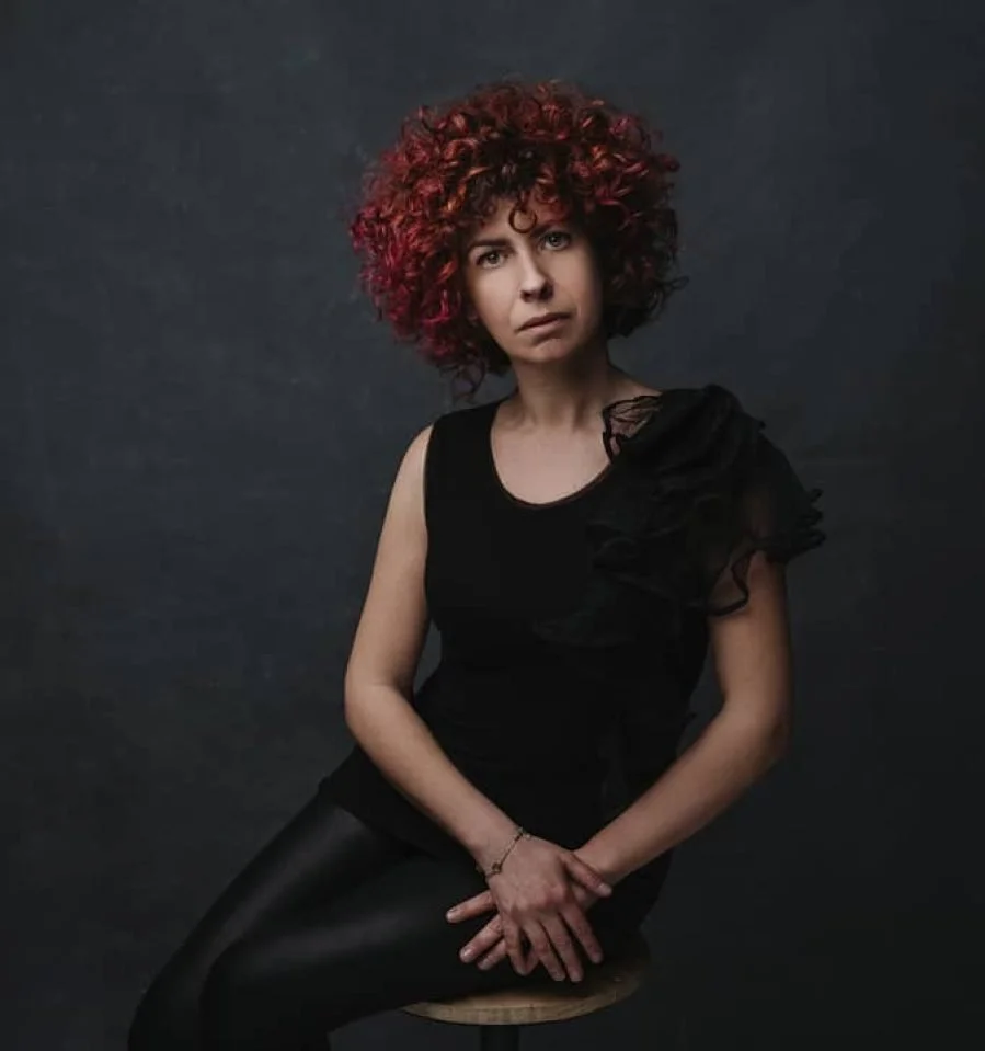 Donna con capelli ricci rossi e vestito nero, seduta su uno sgabello contro sfondo scuro.