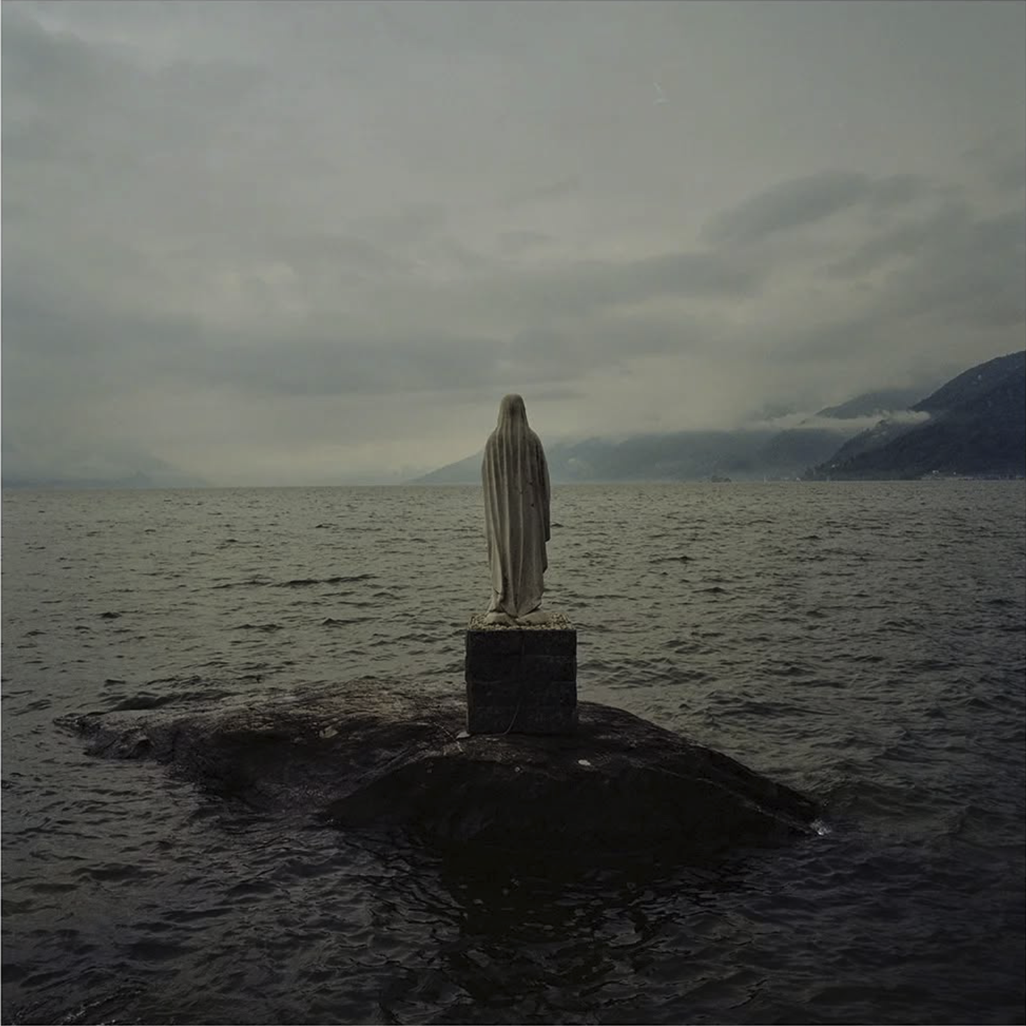 Statua di Gesù Cristo su uno scoglio in mezzo al mare, con montagne e cielo nuvoloso sullo sfondo.