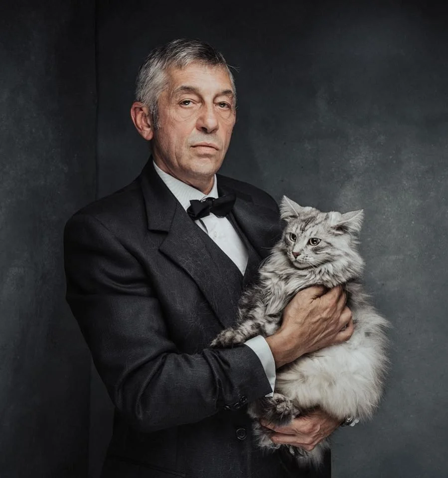 Un uomo con un abito nero elegante e cravatta papillon tiene un gatto a pelo lungo color grigio e bianco.