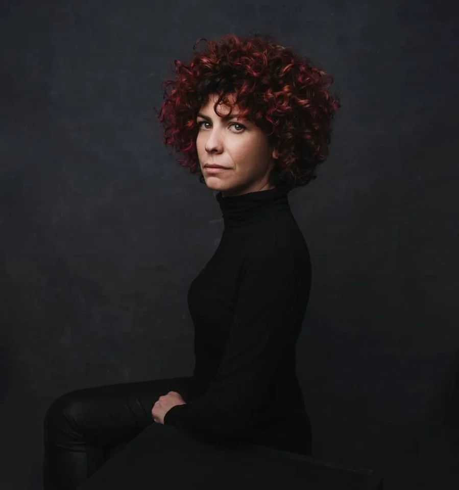 Ragazza con capelli ricciati di colore rosso e neri, vestita con un maglione nero, posa seduta su uno sfondo scuro.
