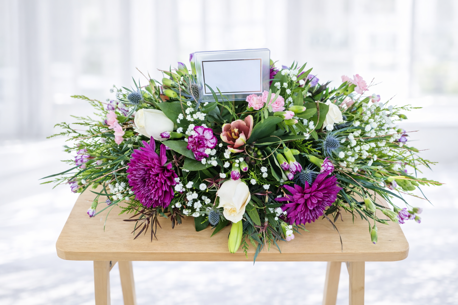 A floral arrangement with purple, pink, white, and green flowers on a wooden table, against a bright background.