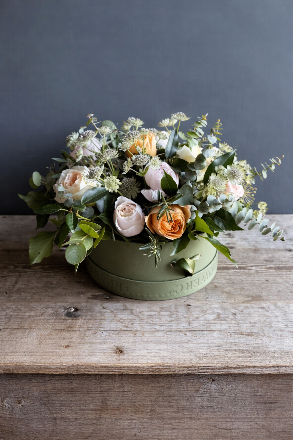 A floral arrangement in a round green box on a wooden surface, featuring roses, eucalyptus leaves, and other greenery against a gray background.