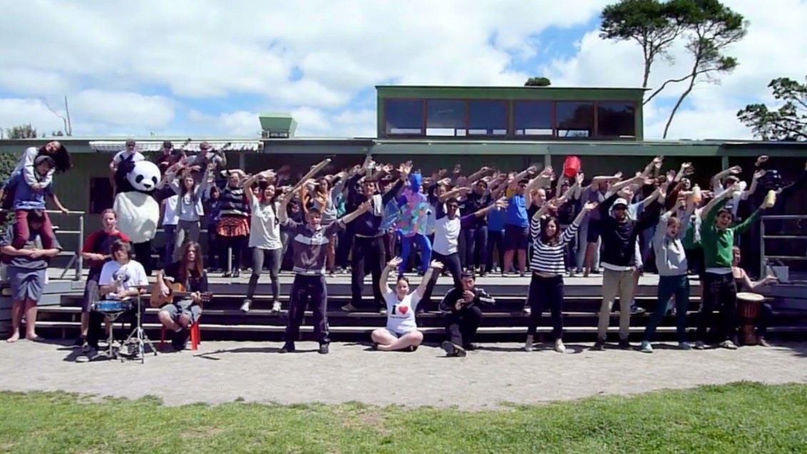 A large group of people, including children and adults, are standing on outdoor steps, raising their arms in a coordinated gesture during a daytime event. Some are dressed casually, and there is a person in a panda costume on the left side. The backg