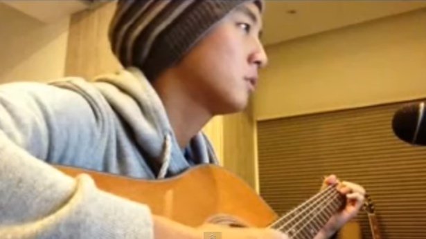 Young man wearing a beanie and gray hoodie playing an acoustic guitar in a room with wooden walls.