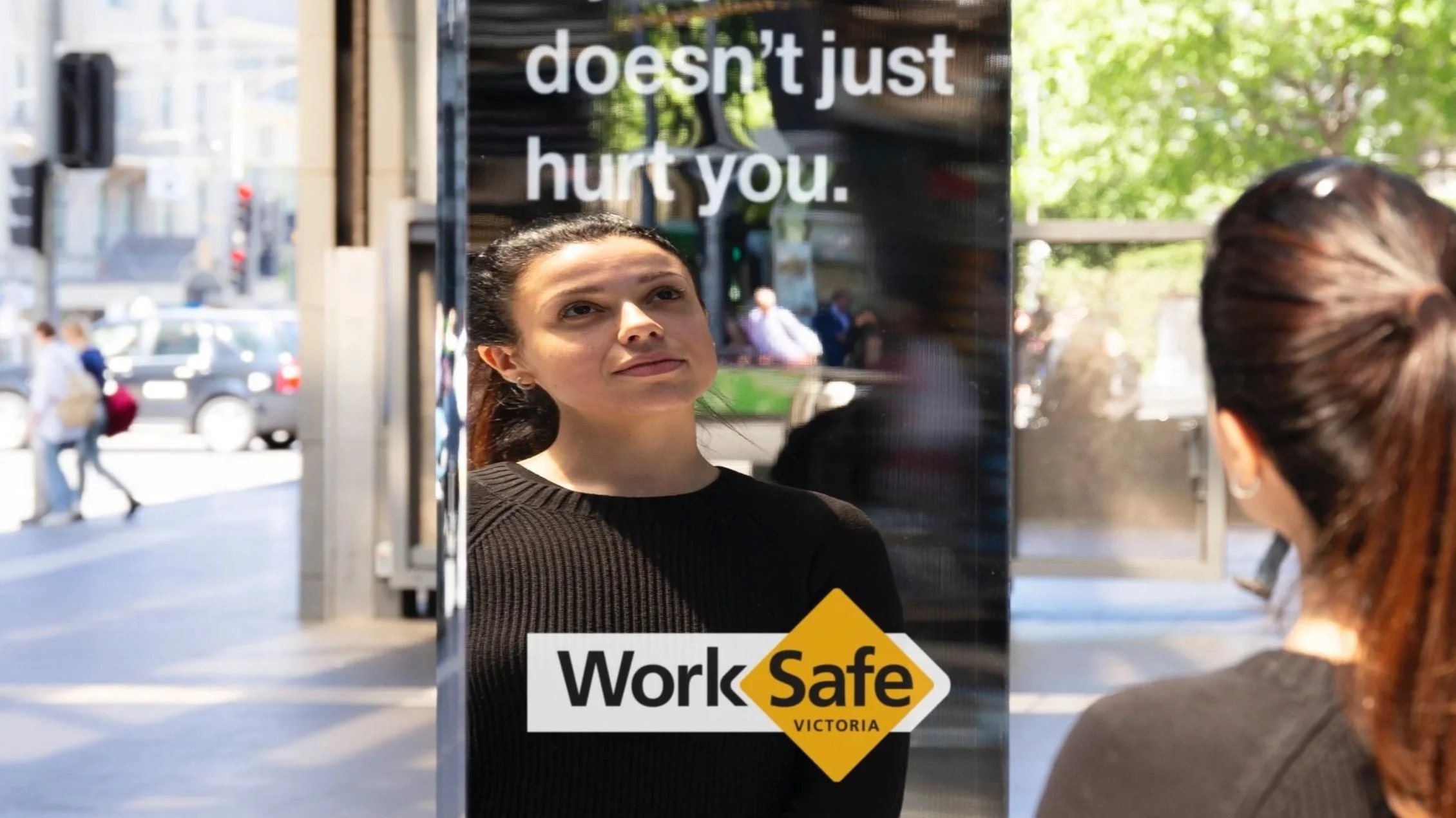 Pat Baron. A woman looks at her reflection in a mirror on a street with a sign that reads 'doesn't just hurt you' and a WorkSafe Victoria sticker.