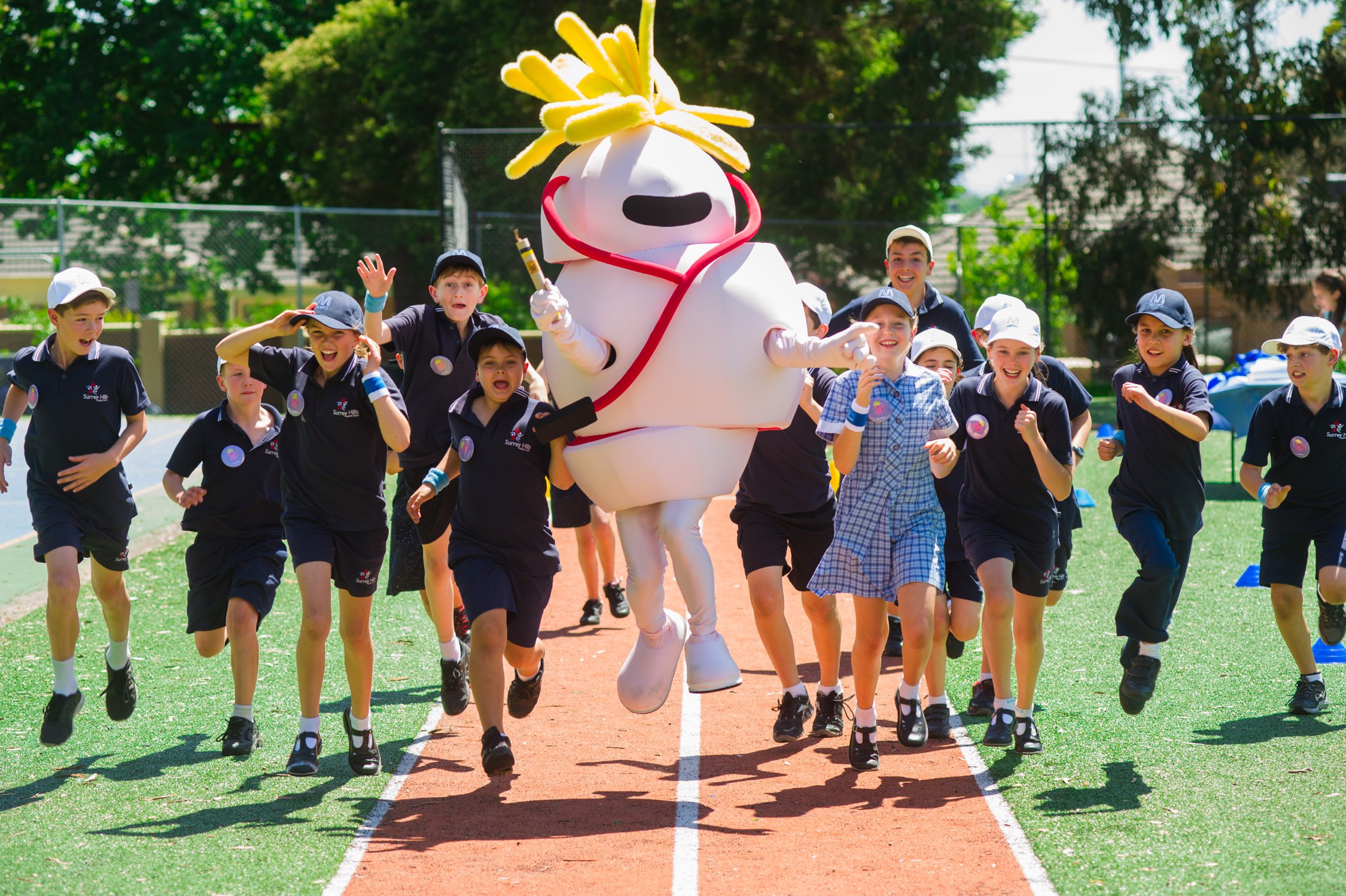 Children running on a track with a large mascot dressed as a nurse with a yellow mop on its head, wearing white scrubs and a stethoscope.