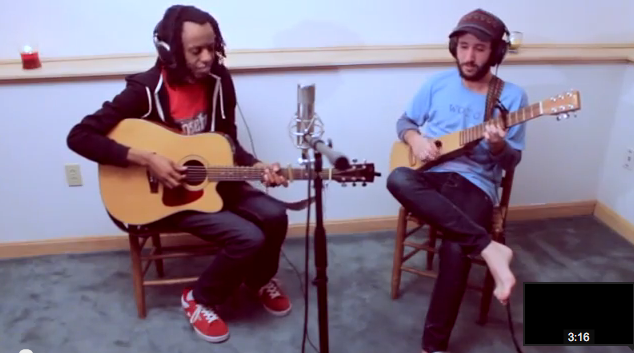 Two men playing acoustic guitars in a recording studio, wearing headphones and sitting on chairs.