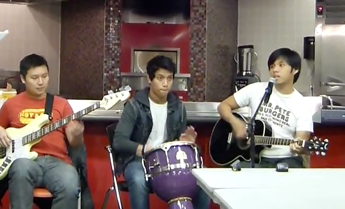 Three young males playing musical instruments: two guitars and a drum, in an indoor setting.