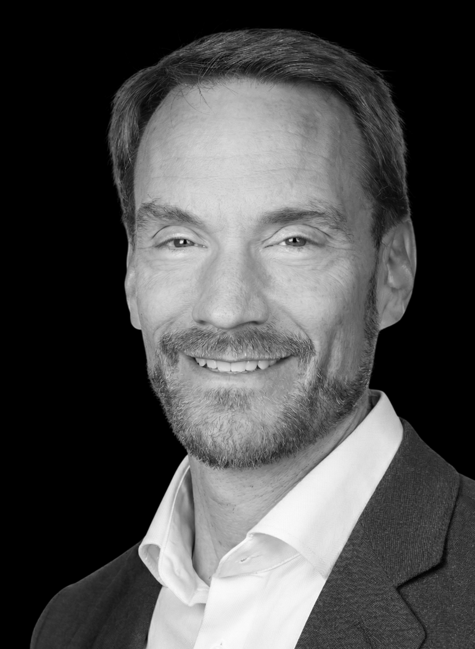 Black and white portrait of a man with short hair and a beard, wearing a checkered button-up shirt, smiling slightly, against a dark background.
