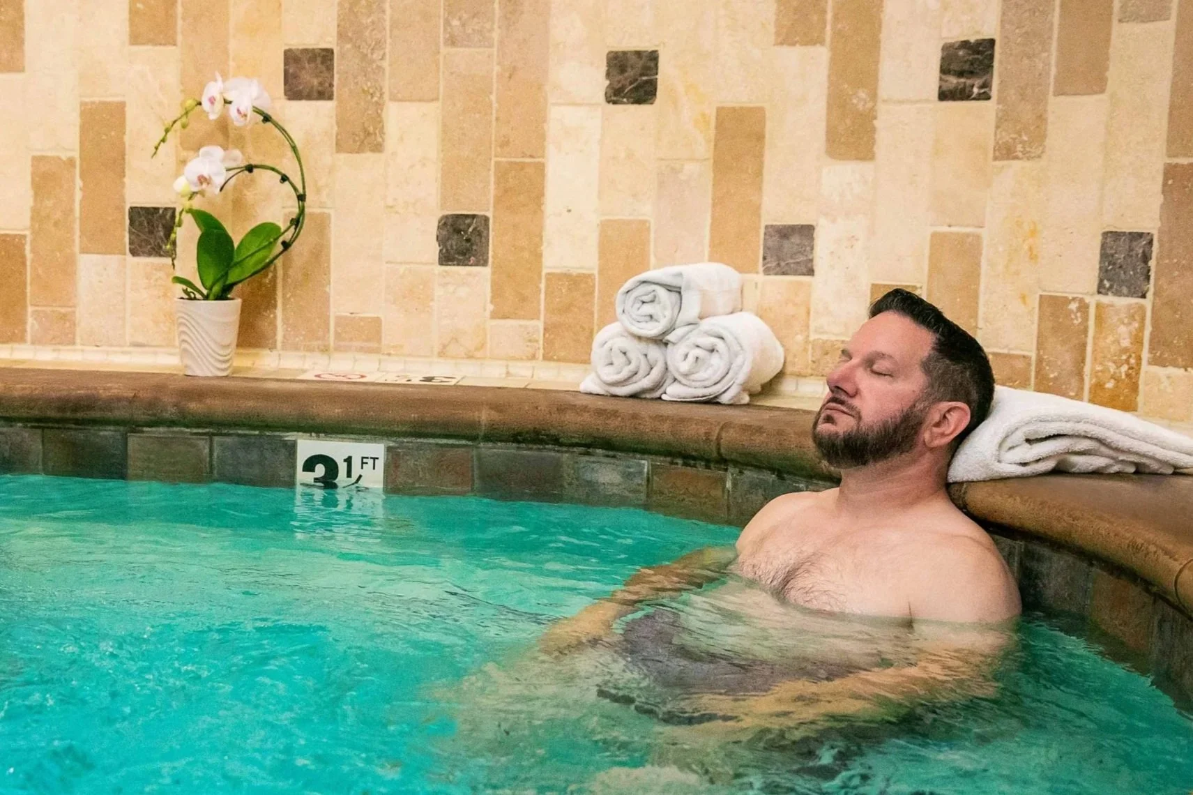 Man relaxing in the whirlpool spa at The Ivy Day Spa, surrounded by warm stone tile and orchids.