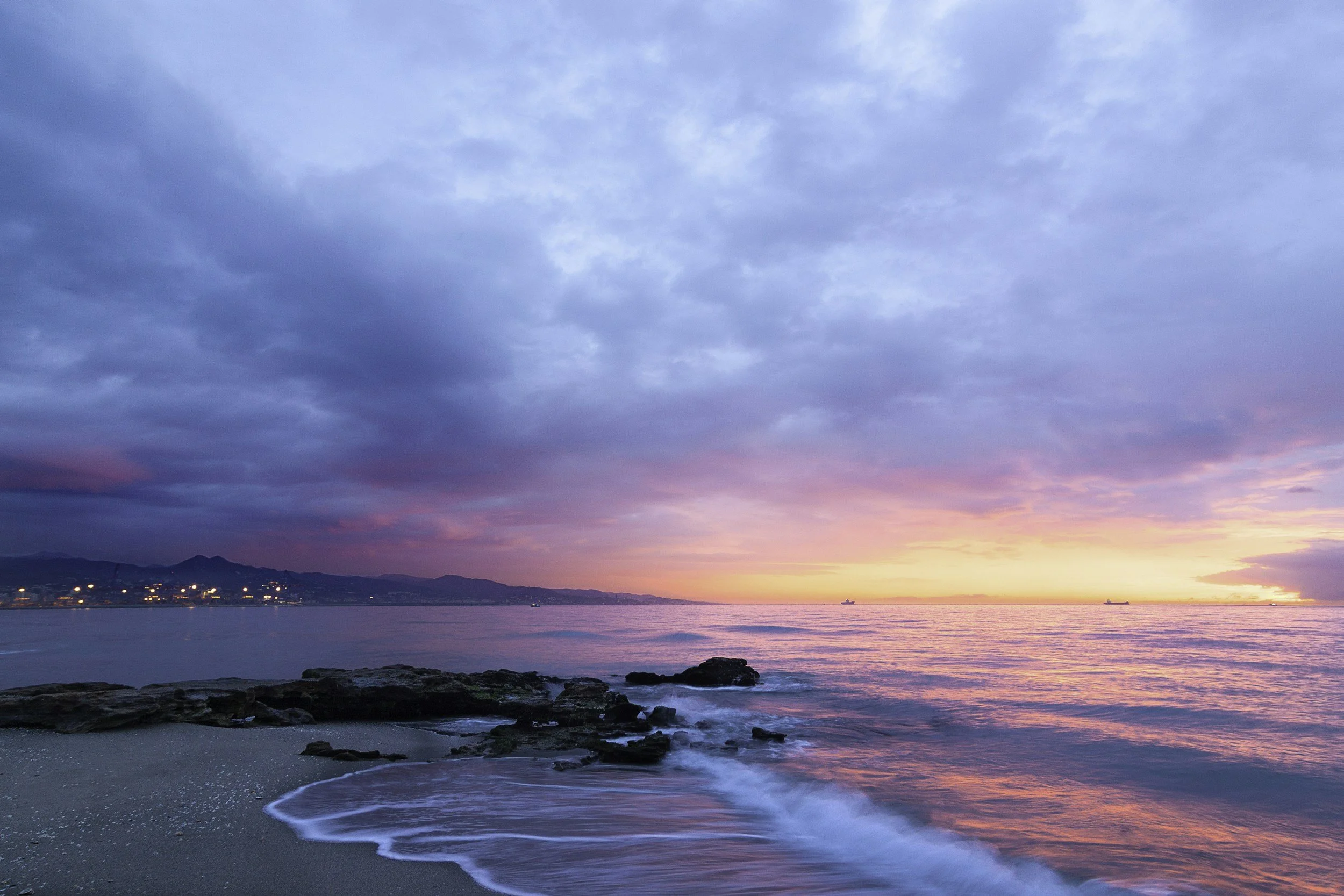 A calm ocean with rocks on the beach in the foreground, a colorful pink and purple sunset, and a partly cloudy sky with mountains and city lights in the distance.