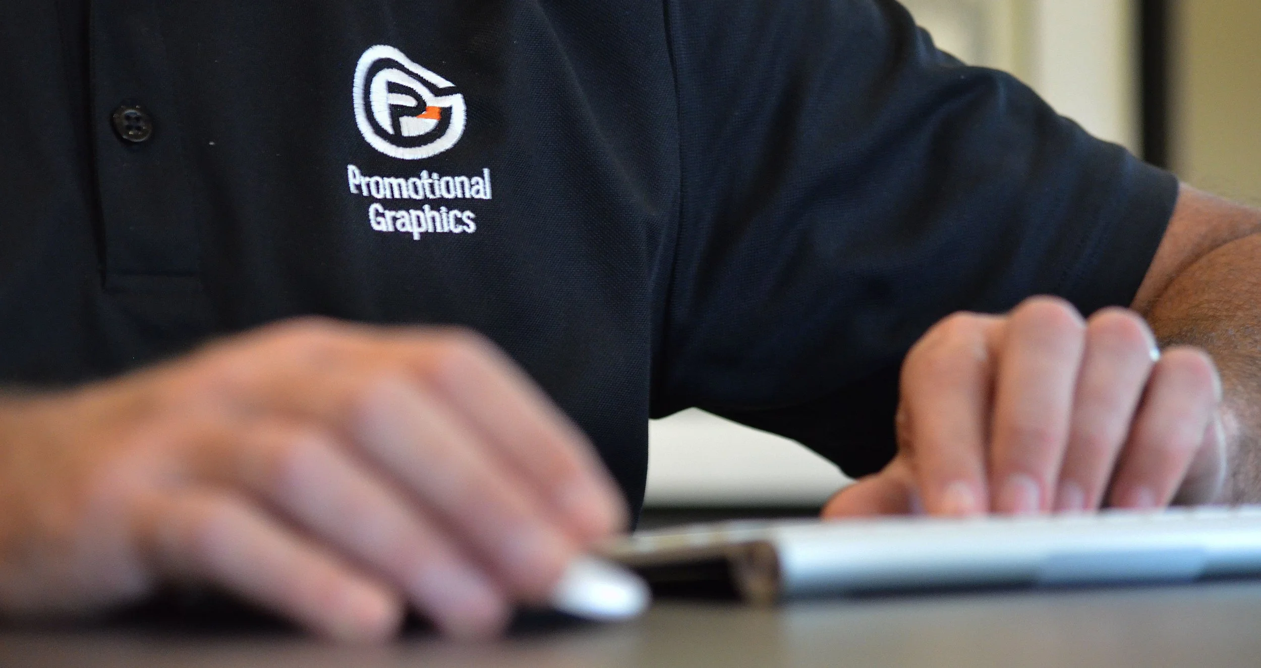 Close-up of a person in a black polo shirt with the logo and text 'Promotional Graphics' on the chest, sitting at a table and interacting with a tablet device.